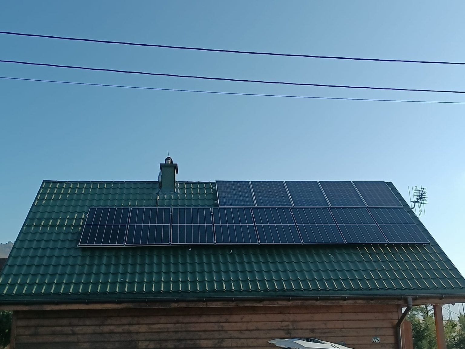 Panele słoneczne na zielonym dachu domu z drewnianą elewacją, antena telewizyjna widoczna po prawej stronie dachu, błękitne niebo w tle.