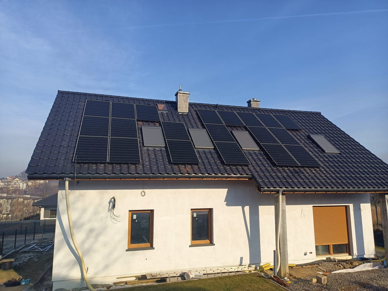 Dom jednorodzinny z panelami słonecznymi na czarnym dachu, widoczne okna dachowe, elewacja w trakcie wykończenia, przewody na ścianie.