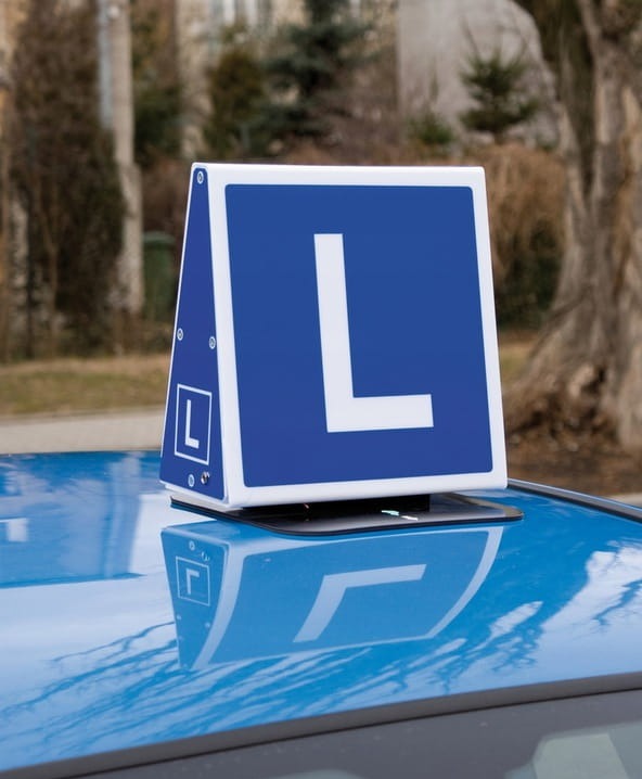 Niebieski samochód z charakterystycznym znakiem 'L' na dachu, oznaczającym naukę jazdy, zaparkowany na tle drzew.