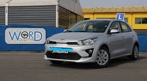 Srebrny samochód Kia Rio z niebieską literą 'L' na dachu, zaparkowany przed szarym budynkiem z niebieskim banerem reklamowym WORD Olsztyn.