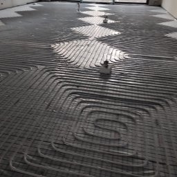 Usługi Instalacje Sanitarne Alicja Zajac - Montaż szarej instalacji ogrzewania podłogowego w surowym pomieszczeniu z widokiem na las przez okno.