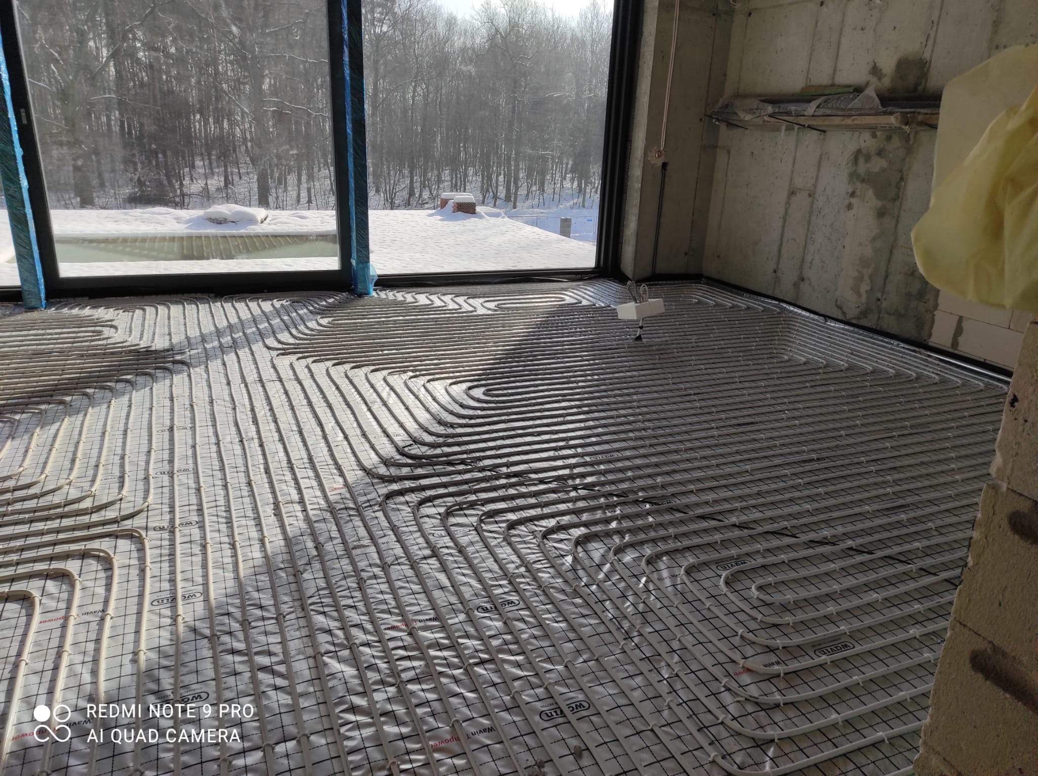Rozłożone rury ogrzewania podłogowego na folii izolacyjnej w surowym wnętrzu z widokiem na zasypany śniegiem las za oknem.