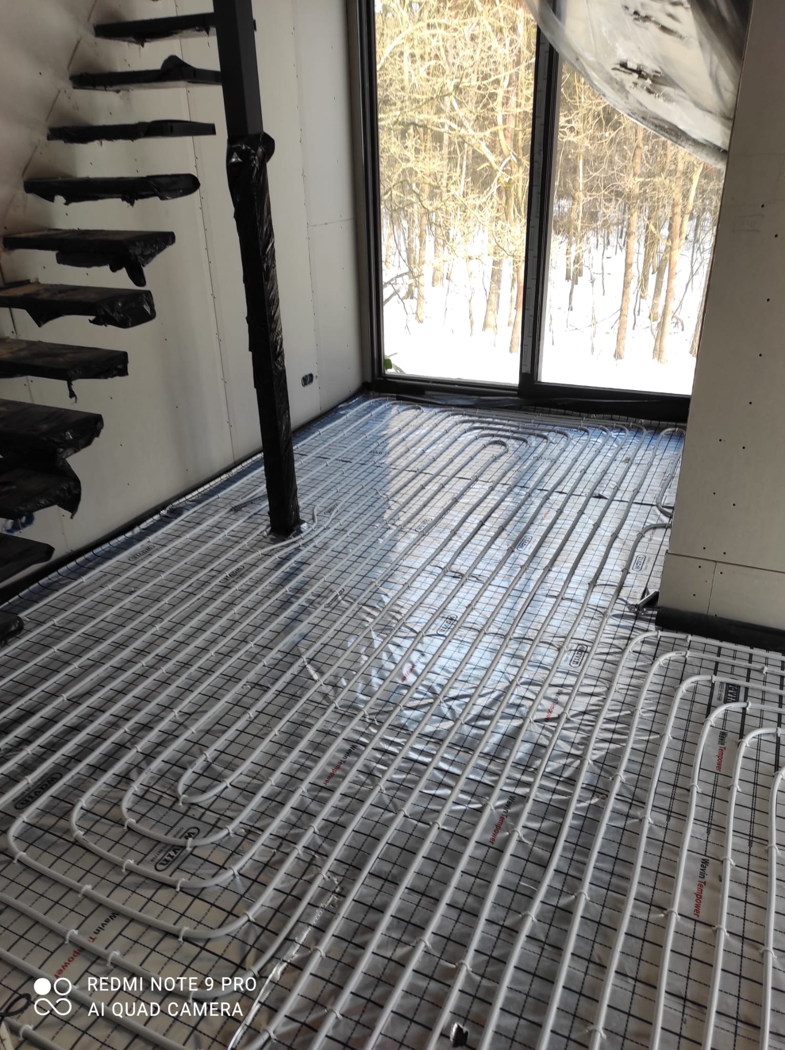 Białe rurki ogrzewania podłogowego ułożone na folii z siatką, widoczne przykryte schody i okno z widokiem na zimowy las.