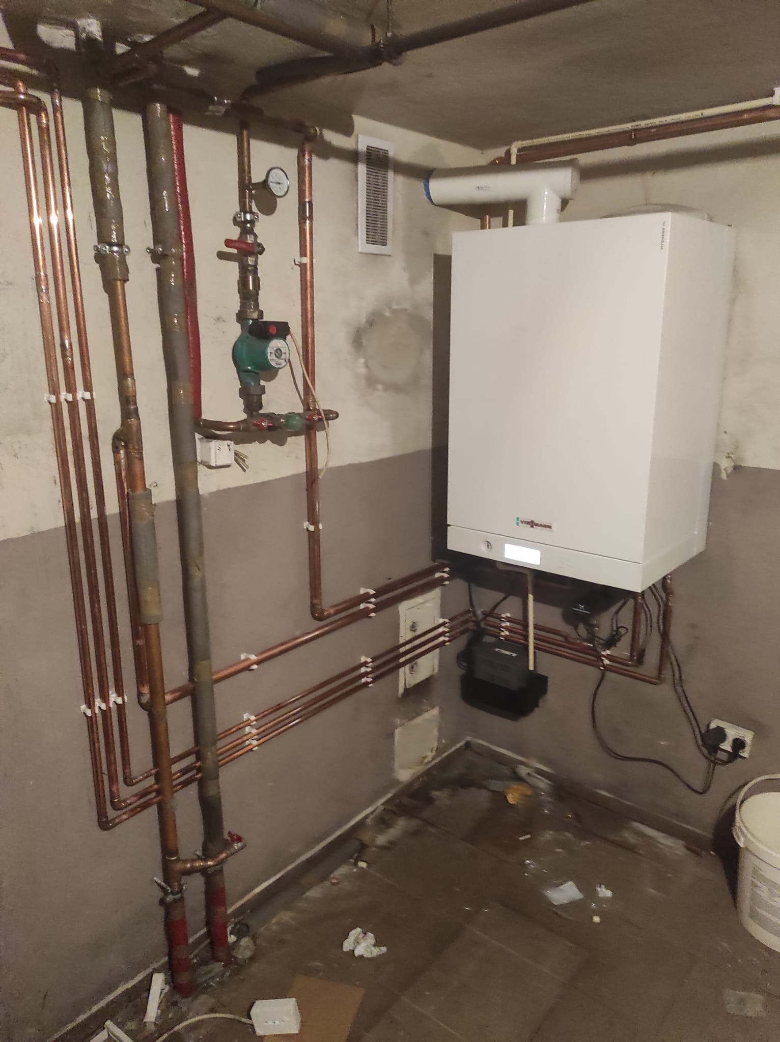 Biały piec gazowy Viessmann zamontowany na ścianie w piwnicy, widoczne miedziane rury instalacji grzewczej, manometr i pompa obiegowa.