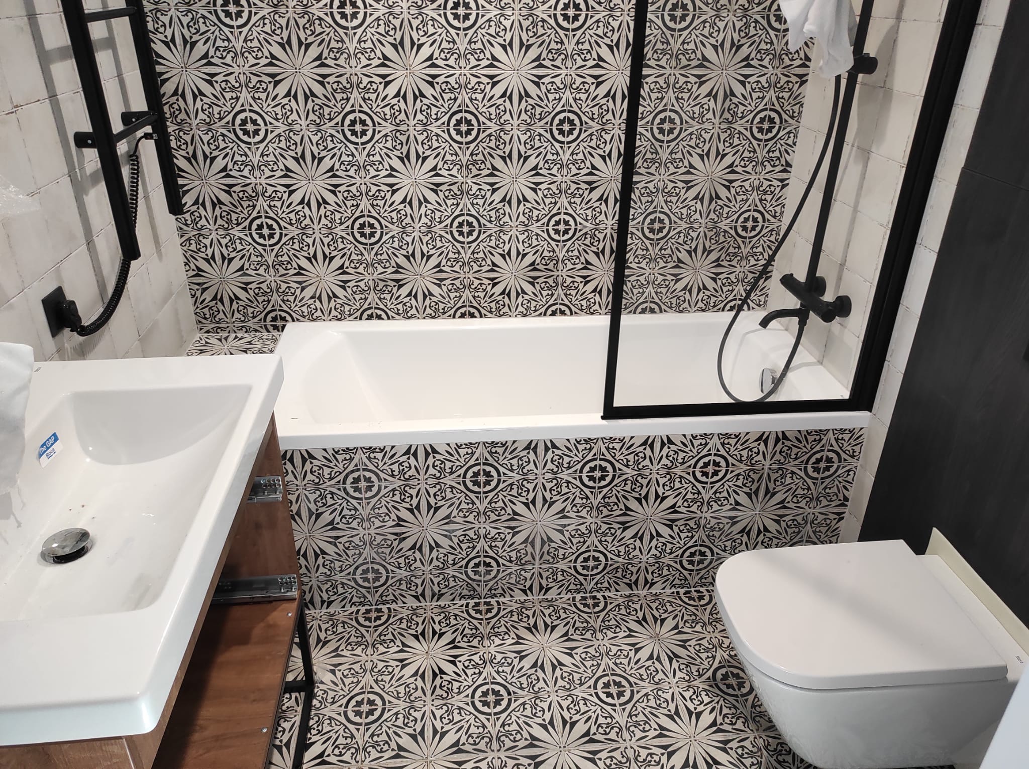 Wykończona łazienka z wanną za czarną szybą, wiszącą toaletą i umywalką Roca Gap na tle wzorzystych czarno-białych płytek podłogowych i ściennych.