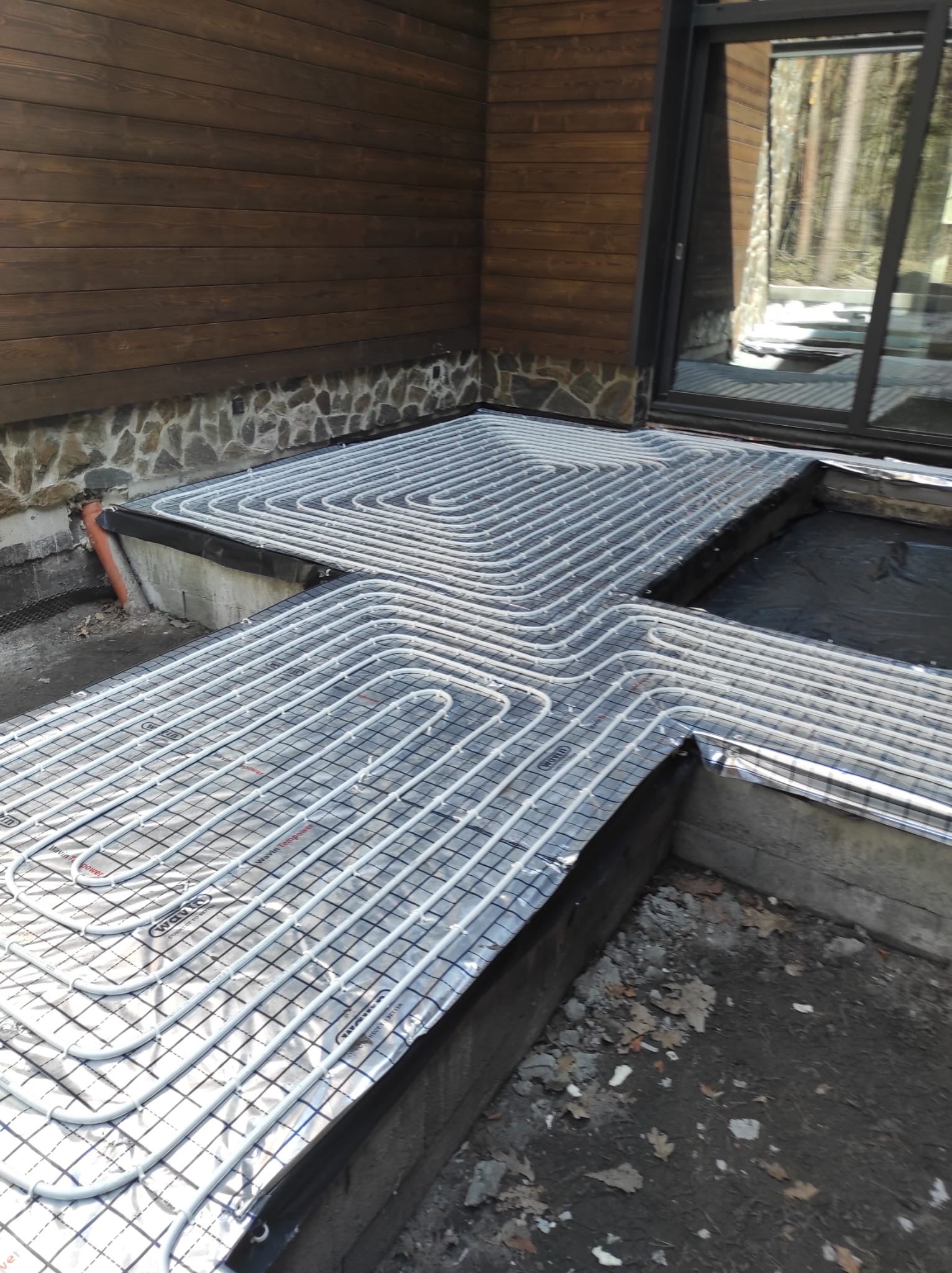 Montaż ogrzewania podłogowego Wavin na folii aluminiowej w zewnętrznej wnęce budynku z drewnianą elewacją i kamienną podmurówką, widok na okno tarasowe.