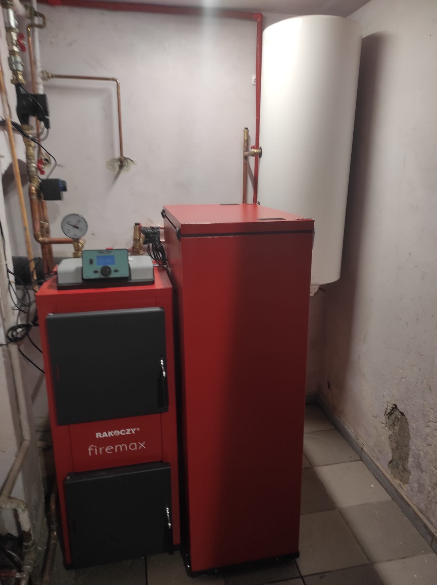 Czerwony piec grzewczy RAKOCZY firemax z podłączonymi rurami miedzianymi i sterownikiem elektronicznym, obok biały zasobnik ciepłej wody, w tle ściana z widocznymi instalacjami.