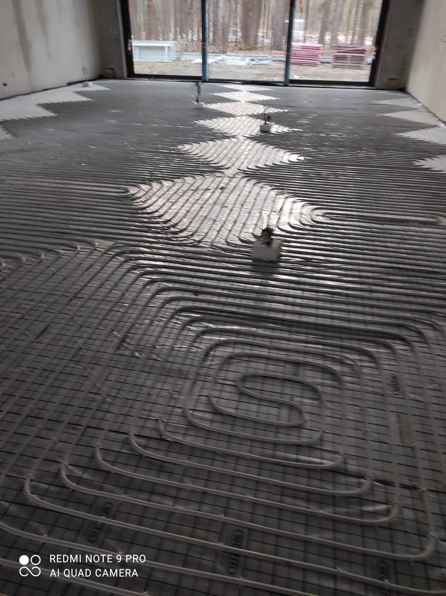 Montaż szarej instalacji ogrzewania podłogowego w surowym pomieszczeniu z widokiem na las przez okno.