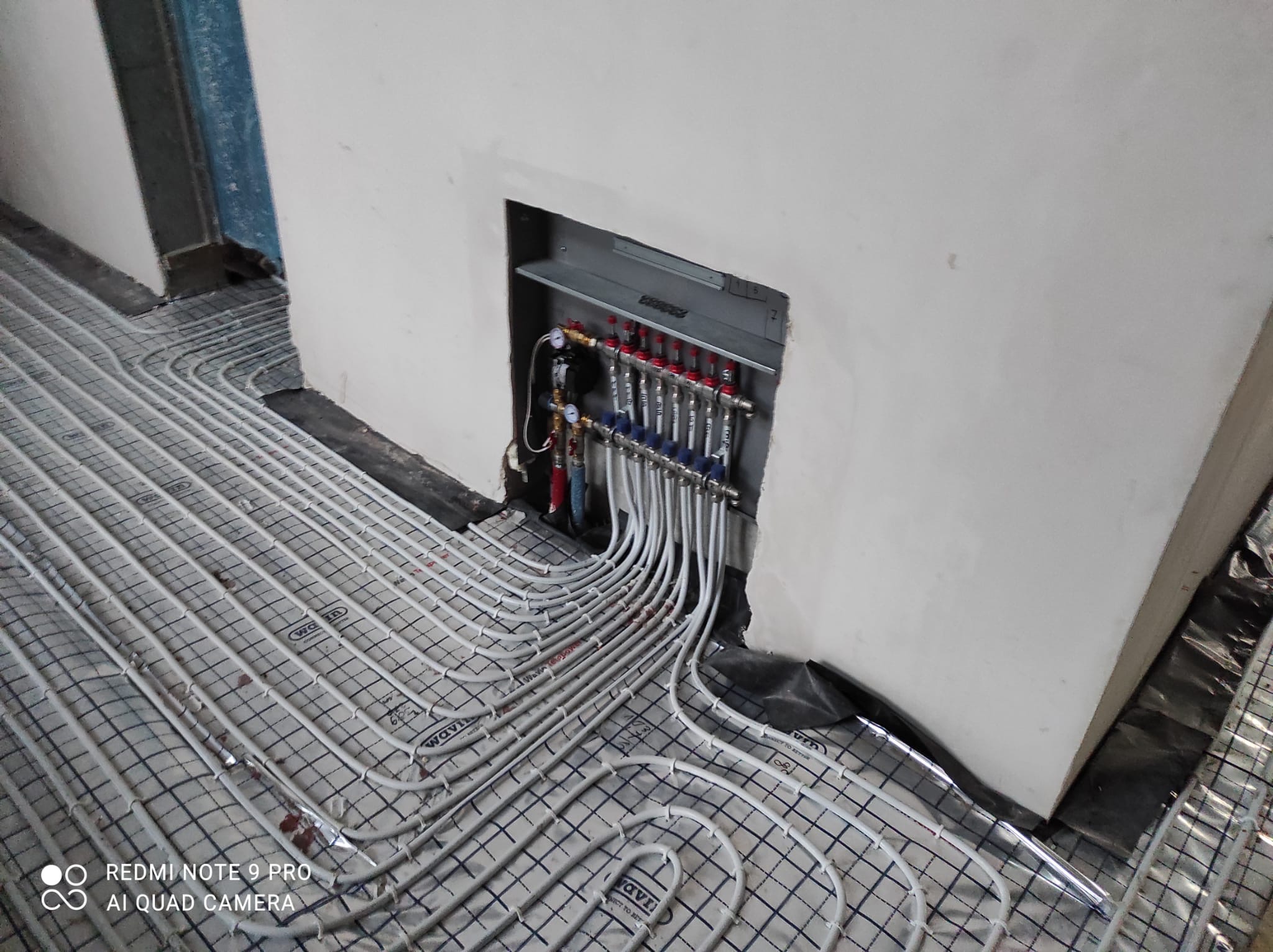 Rozłożone rury ogrzewania podłogowego na izolacji z widocznym rozdzielaczem w ścianie, rury ułożone w pętle, widoczne elementy montażowe i izolacja przy ścianach.