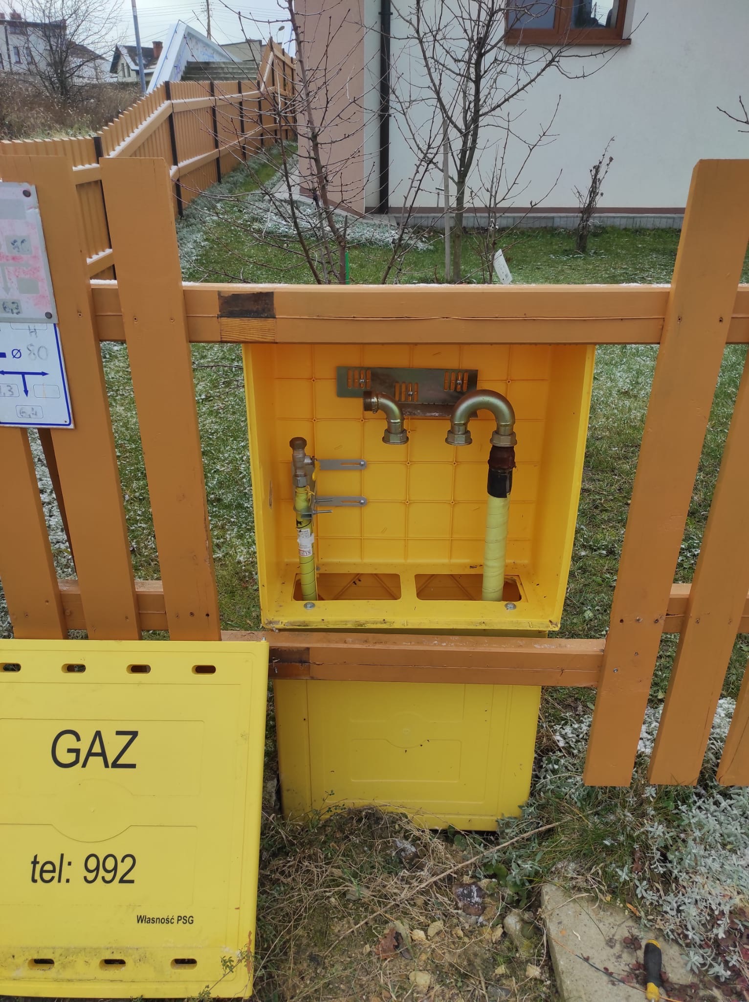 Żółta skrzynka gazowa z widocznymi przyłączami, umieszczona w drewnianym ogrodzeniu na zewnątrz budynku w zimowej scenerii.