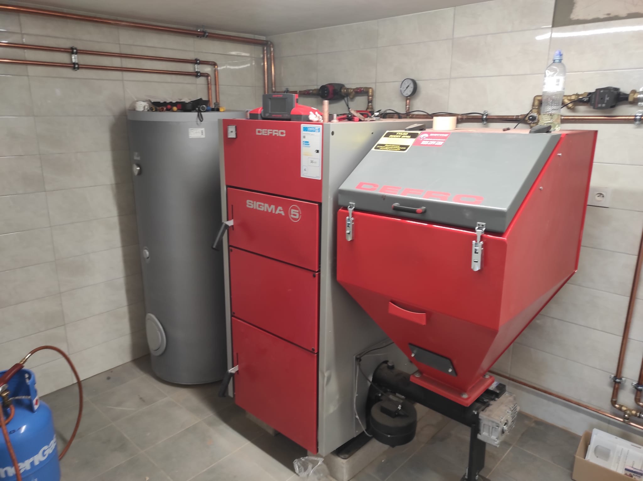 Czerwono-szary piec CO marki Defro Sigma 5 z zasobnikiem, zbiornik na wodę, instalacja z miedzianych rur na ścianie z płytek, butla gazowa w rogu pomieszczenia.