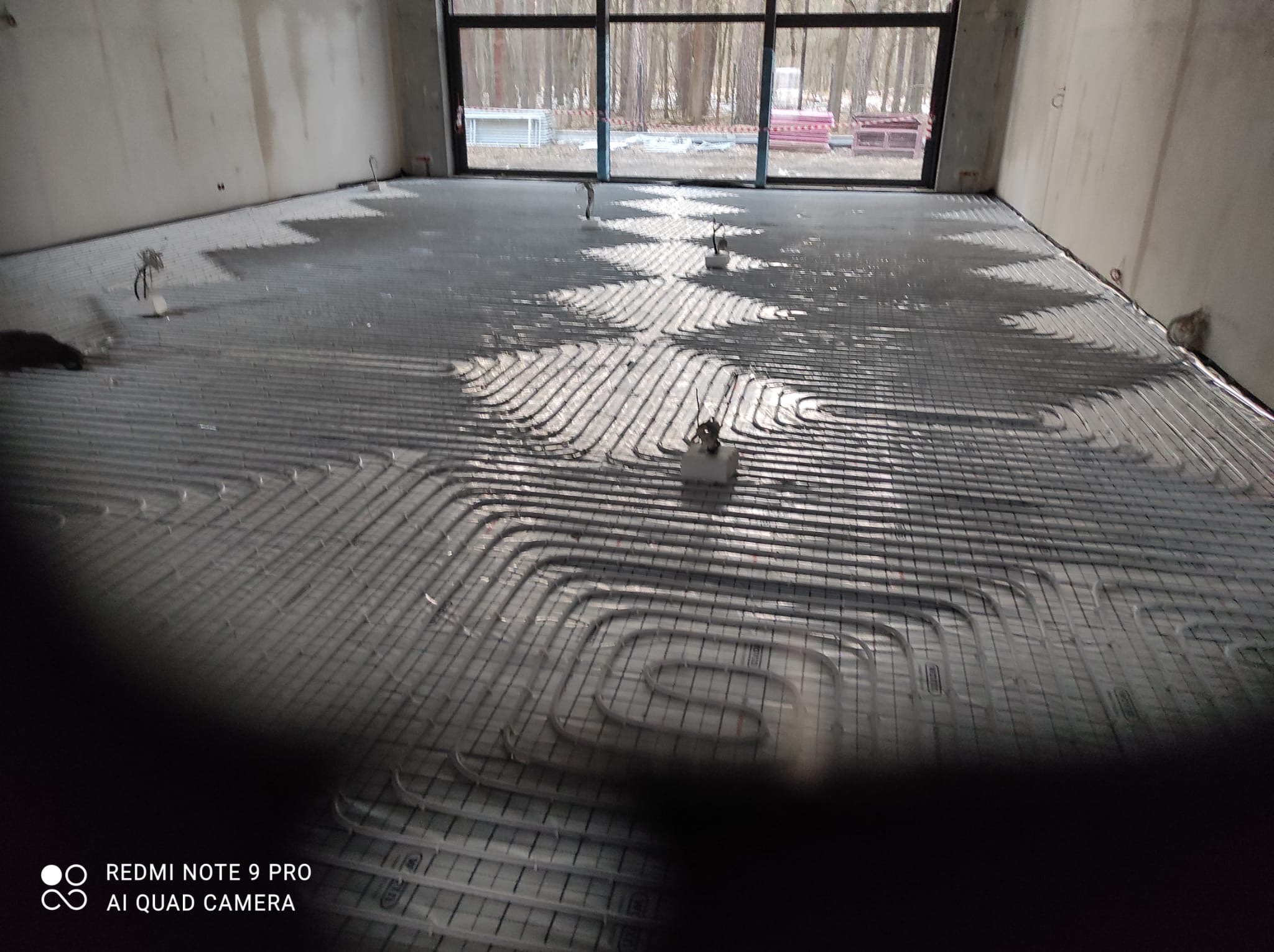 Instalacja ogrzewania podłogowego w surowym stanie, widoczne rury ułożone na podłodze przed wylewką, duże okno z widokiem na drzewa w tle.