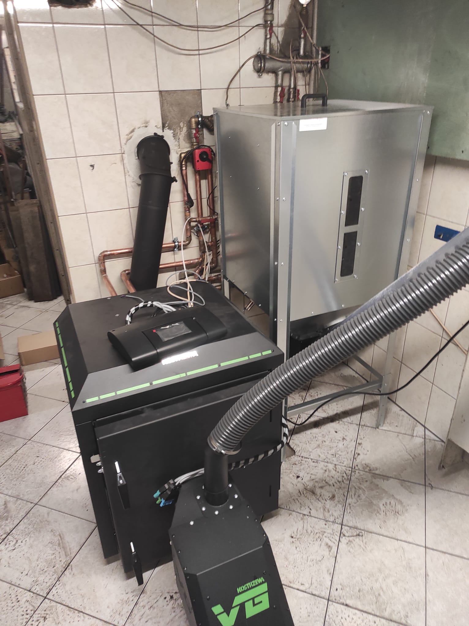 Kotłownia z zamontowanym kotłem na paliwo stałe marki Kostrzewa VG, połączonym z zasobnikiem i instalacją centralnego ogrzewania w pomieszczeniu z wykończeniem w płytkach ceramicznych.