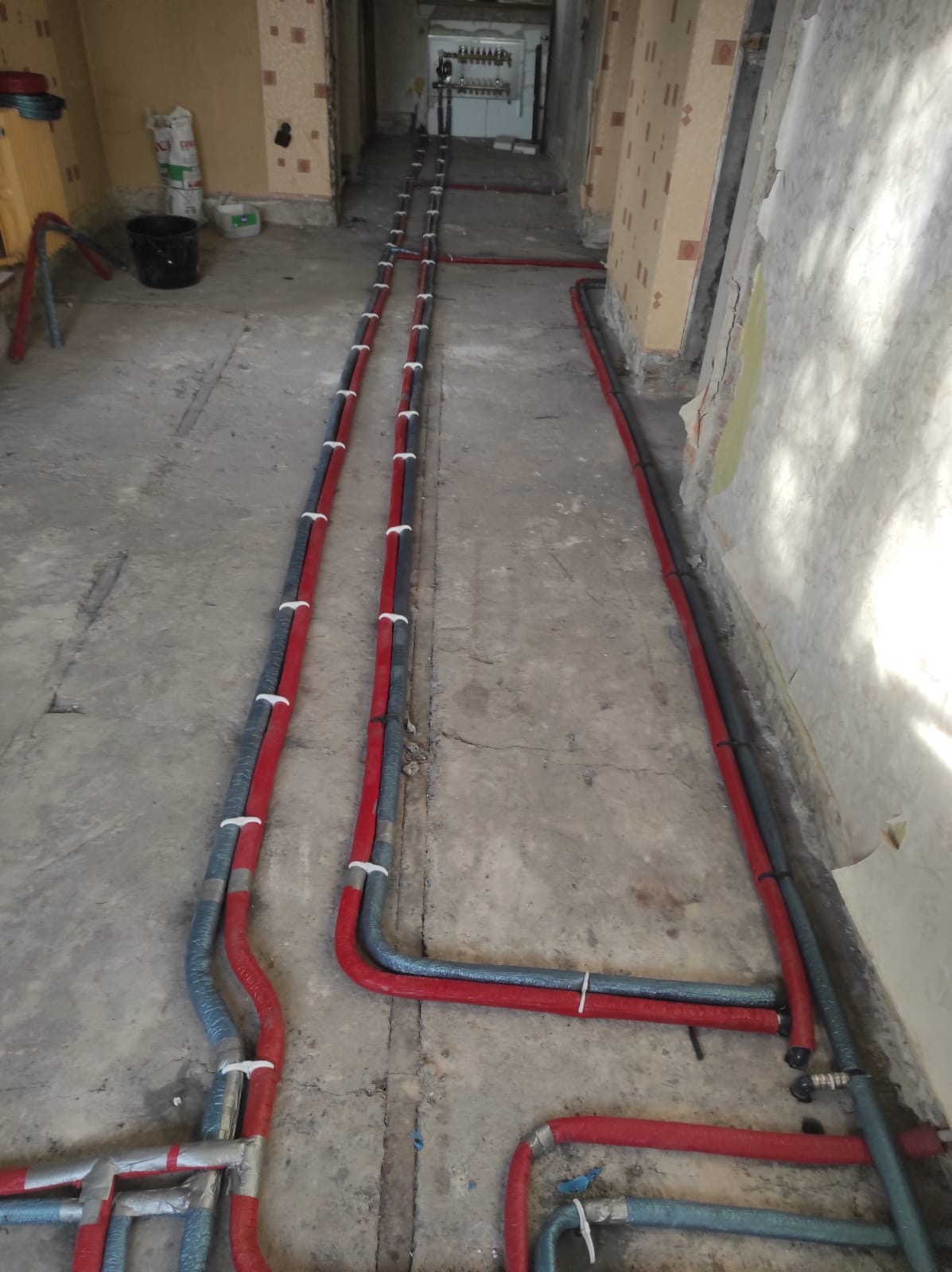 Czerwono-szare rury ogrzewania podłogowego, zabezpieczone białymi opaskami, ułożone wzdłuż korytarza w trakcie remontu.