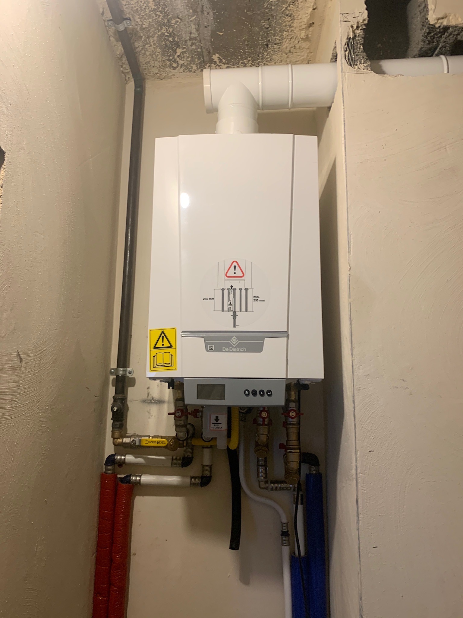 Biały, wiszący piec gazowy De Dietrich z podłączonymi rurami miedzianymi i plastikowymi w surowym pomieszczeniu z widocznymi śladami po kuciu w ścianie.