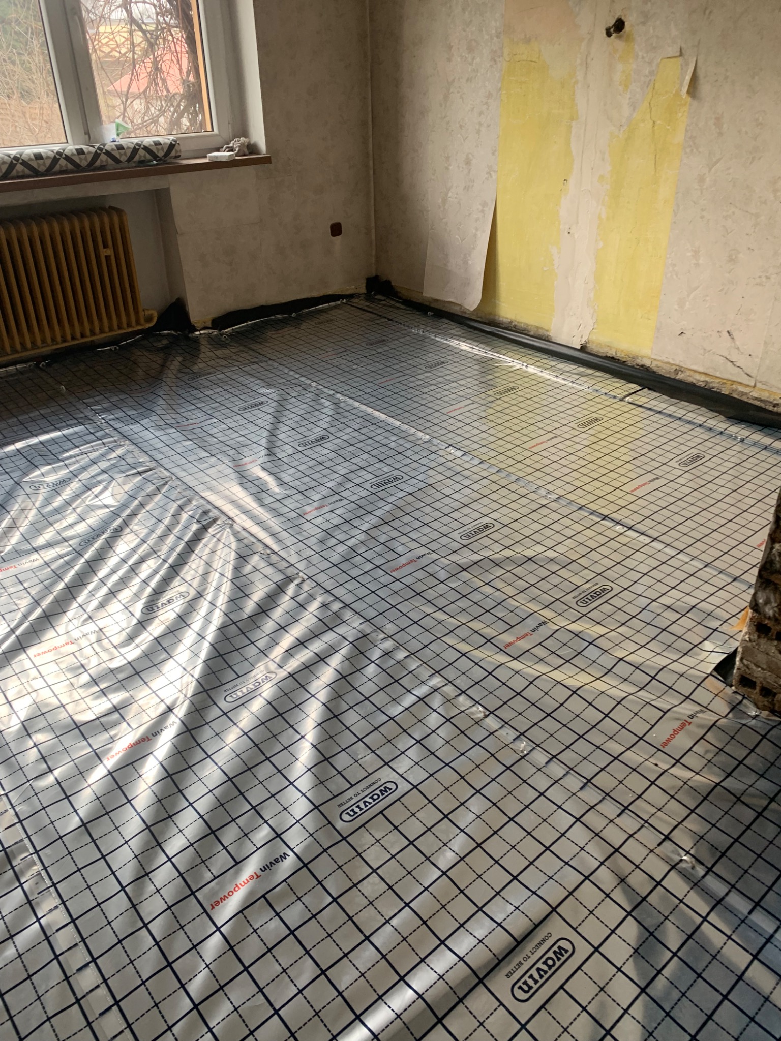 Przygotowanie podłoża pod instalację ogrzewania podłogowego: folia systemowa z nadrukowaną siatką, widoczny fragment ściany z resztkami tapety i stary grzejnik żeberkowy.