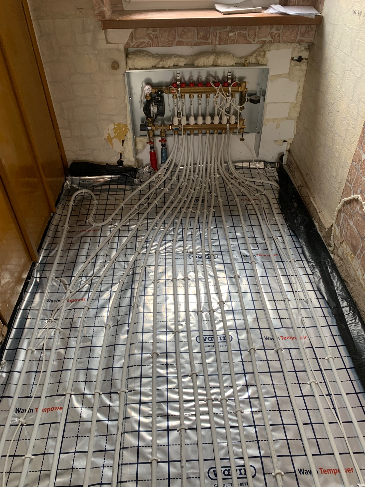 Rozłożona instalacja ogrzewania podłogowego Wavin na folii termoizolacyjnej z widocznym rozdzielaczem i rurami wychodzącymi z niego, w surowym stanie wykończenia wnętrza.