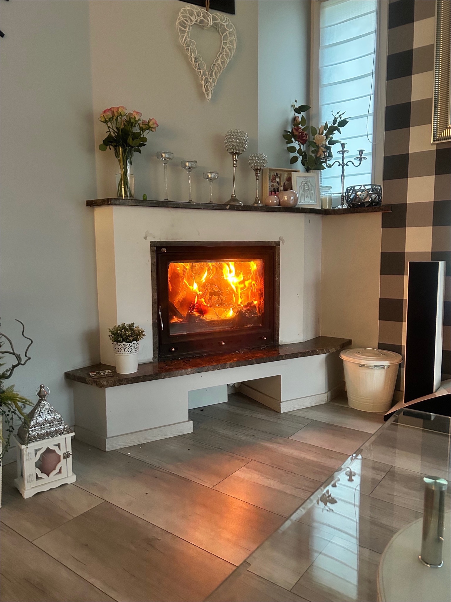 Otwarty kominek z płomieniami w salonie, dekoracje na kominku, wzorzysta tapeta w tle, odbicie płomieni na szklanym stoliku.