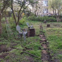 Szybki ogród - Wykopany rów w trawie, przygotowany pod ścieżkę z kostki, obok leżą sterty kostki i dwa plastikowe krzesła, w tle drzewa owocowe i budynek mieszkalny.