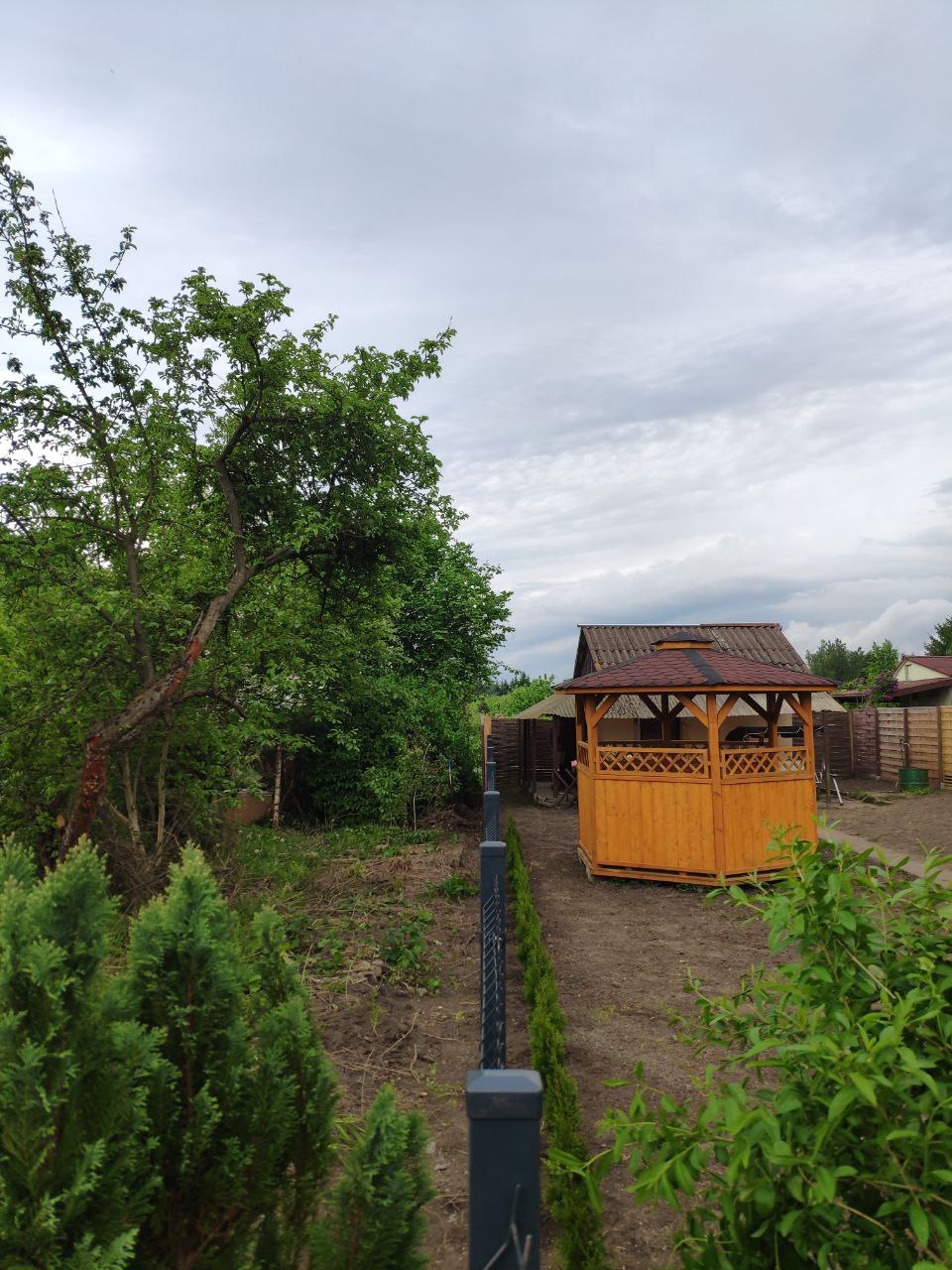 Nowoczesne ogrodzenie panelowe w kolorze grafitowym oddzielające trawnik od ścieżki z drewnianą altaną w tle, otoczone zielenią w pochmurny dzień.