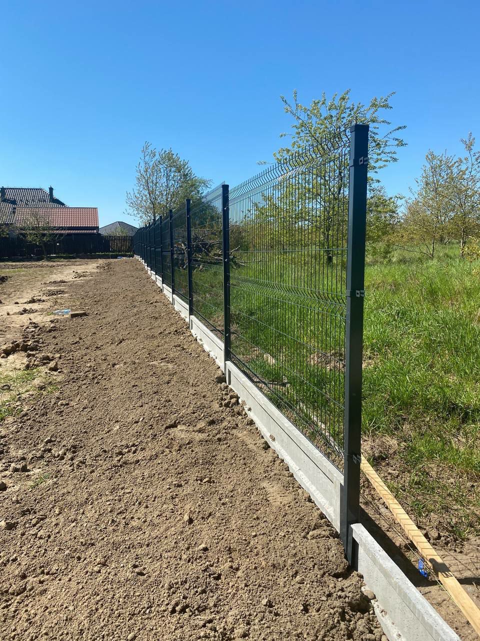 Nowo postawione, panelowe ogrodzenie z podmurówką betonową na świeżo wyrównanym terenie, widok z boku, słoneczny dzień.