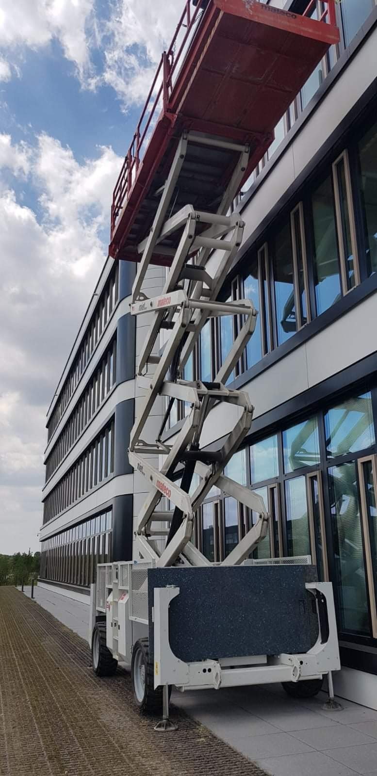 Podnośnik koszowy Mateco ustawiony przy nowoczesnym biurowcu z licznymi oknami, gotowy do prac na wysokości.