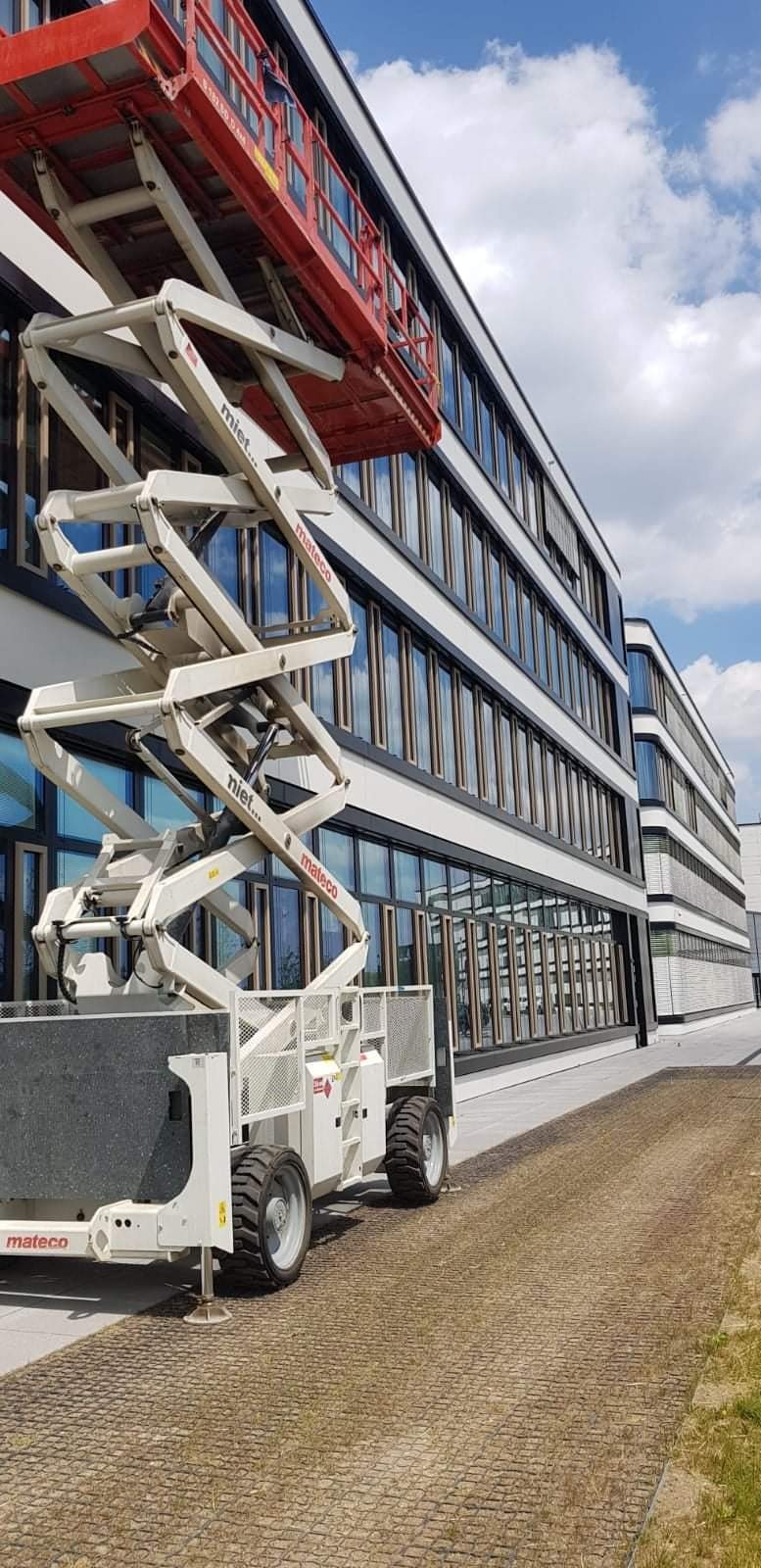 Podnośnik koszowy Mateco przed budynkiem biurowym z licznymi oknami, widoczne odbicia chmur w szybach.