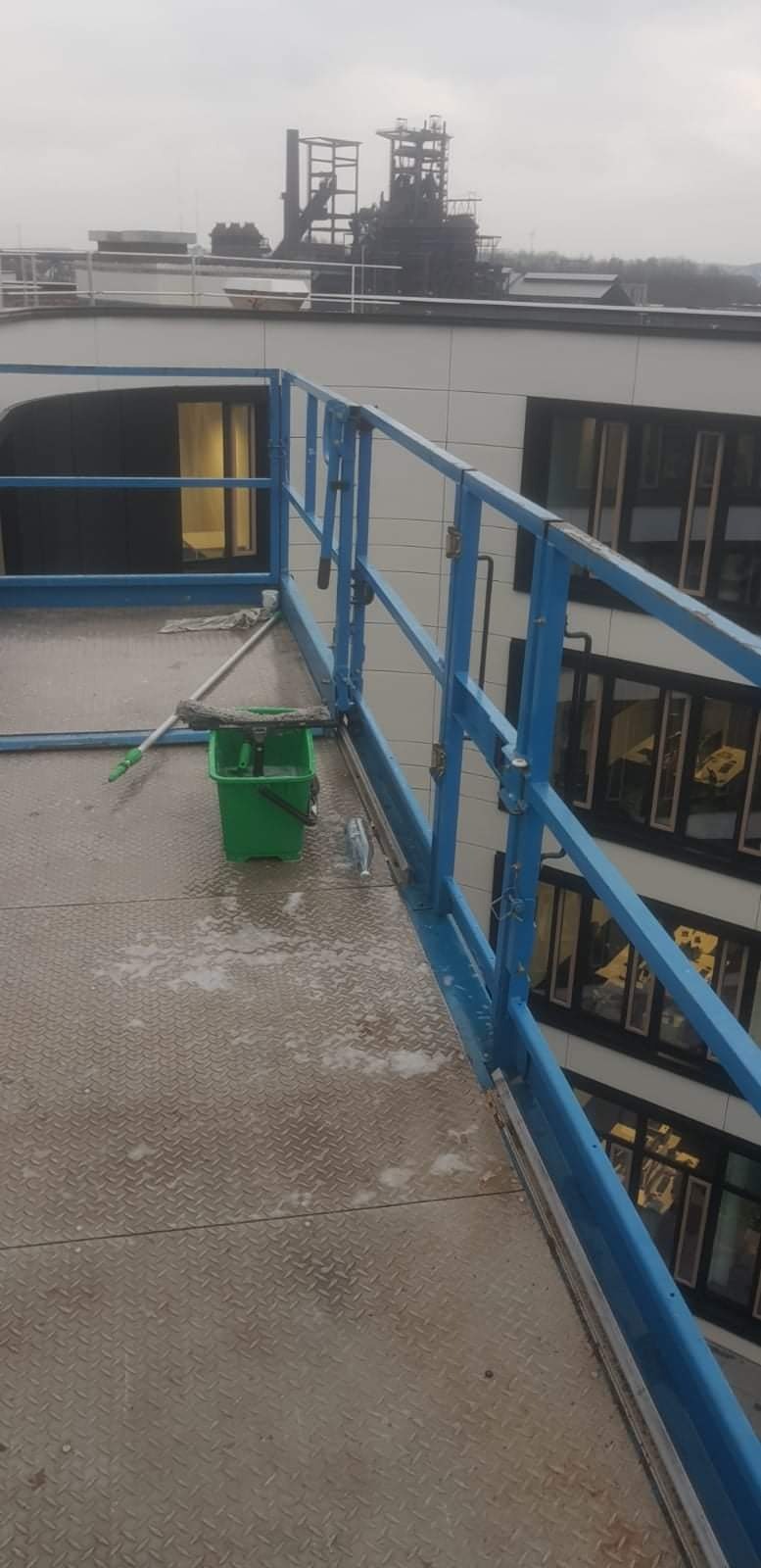 Widok z wysokości na platformę roboczą z niebieskimi barierkami, zielonym wiadrem z mopem i myjką do okien, na podłożu pozostałości po myciu, w tle budynek z oknami i konstrukcja przemysłowa.