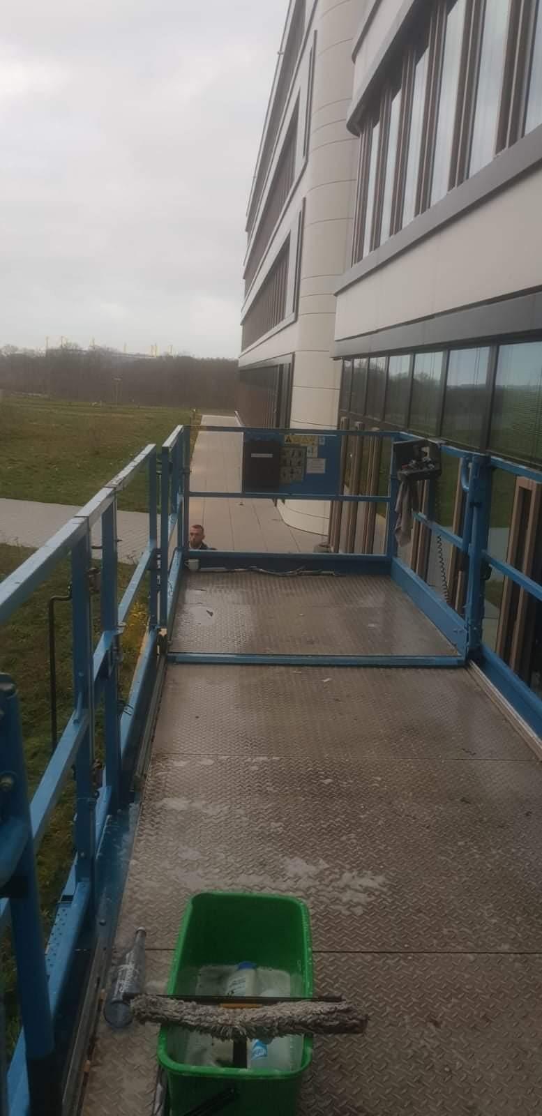Widok z podnośnika koszowego na elewację biurowca w trakcie prac, widoczne narzędzia i wiadro z płynem do mycia, pracownik w tle.