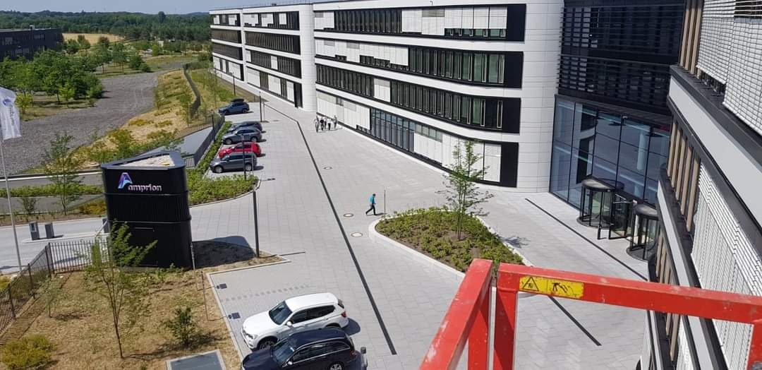 Nowoczesny biurowiec z szarymi oknami i elewacją w odcieniach bieli, widok z wysokości z elementami czerwonego podnośnika w prawym dolnym rogu, na pierwszym planie parking z zaparkowanymi...