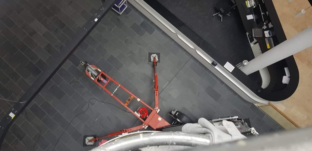 Widok z góry na maszynę do mycia okien na wysokości, przymocowaną przyssawkami do ciemnej, kafelkowanej powierzchni, z fragmentem białej rury i biurkiem w tle.