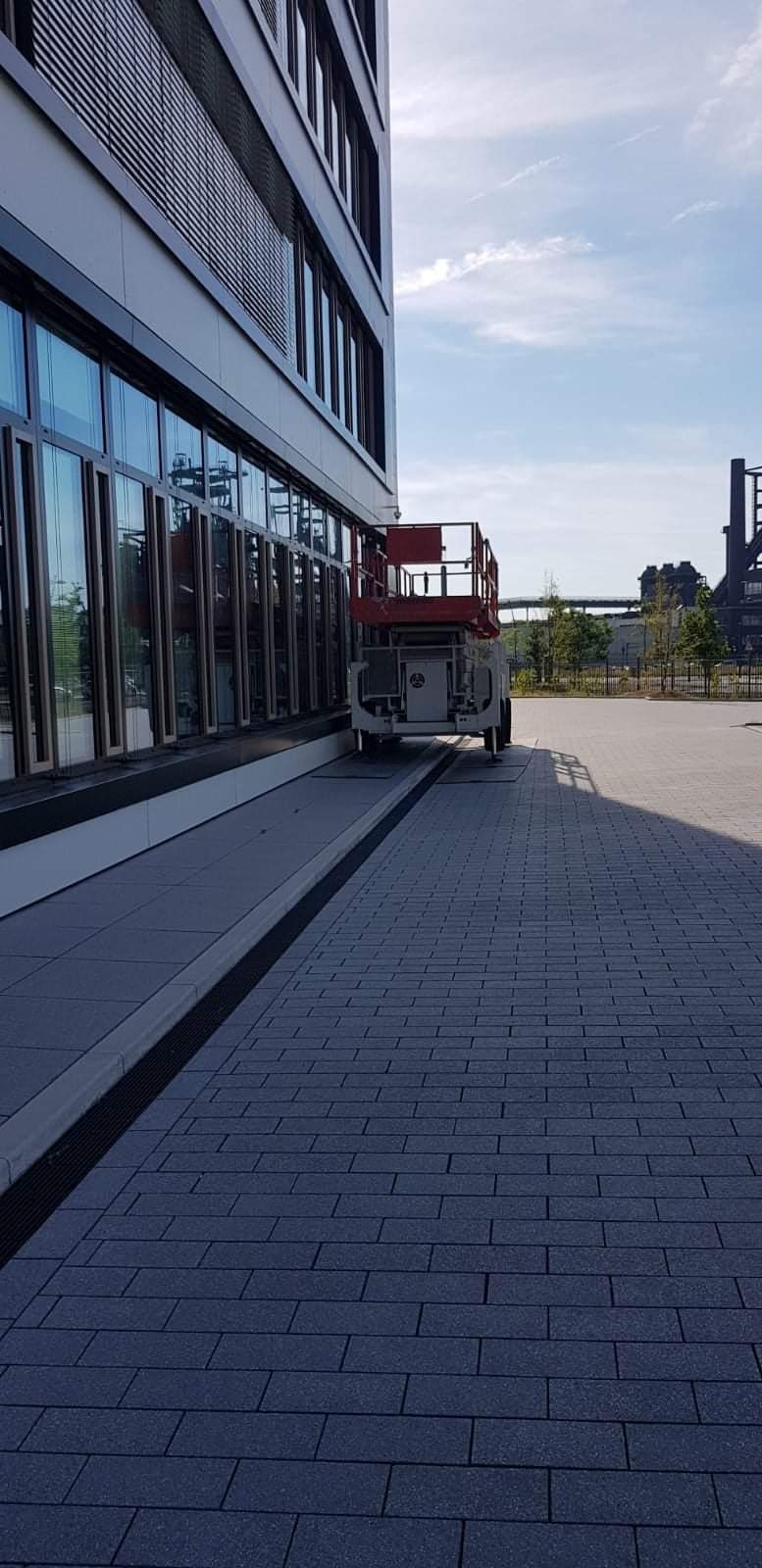 Podnośnik koszowy zaparkowany przy szklanej fasadzie nowoczesnego biurowca na tle przemysłowego krajobrazu.