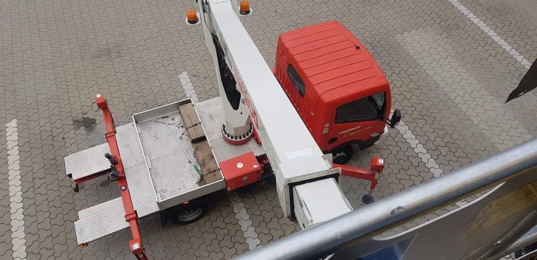 Widok z góry na czerwono-biały podnośnik koszowy na brukowanym placu, z widocznym koszem i elementami zabezpieczającymi.