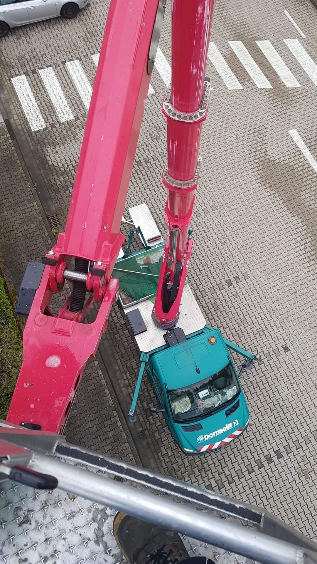 Widok z góry na podnośnik koszowy z wysięgnikiem w kolorze różowym, zaparkowany na chodniku przy przejściu dla pieszych, obok zielonego samochodu dostawczego z logo firmy Domselff.