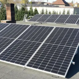 Panele słoneczne zamontowane na płaskim dachu budynku mieszkalnego, widoczne kominy i drabina oparta o dach.