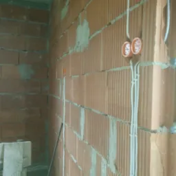 Surowe ściany z cegły dziurawki z widocznymi puszkami elektrycznymi i przewodami zabezpieczonymi uchwytami, w trakcie prac instalacyjnych.