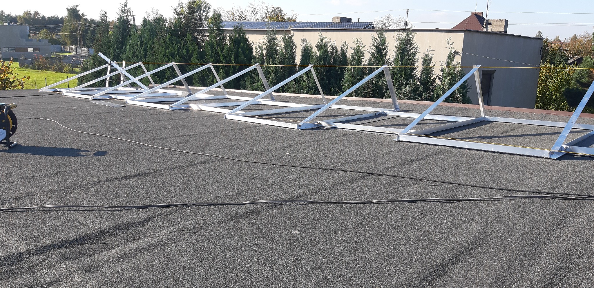 Metalowe konstrukcje wsporcze do paneli fotowoltaicznych, ustawione rzędami na płaskim, bitumicznym dachu budynku, z widocznym przewodem elektrycznym i budynkami w tle.