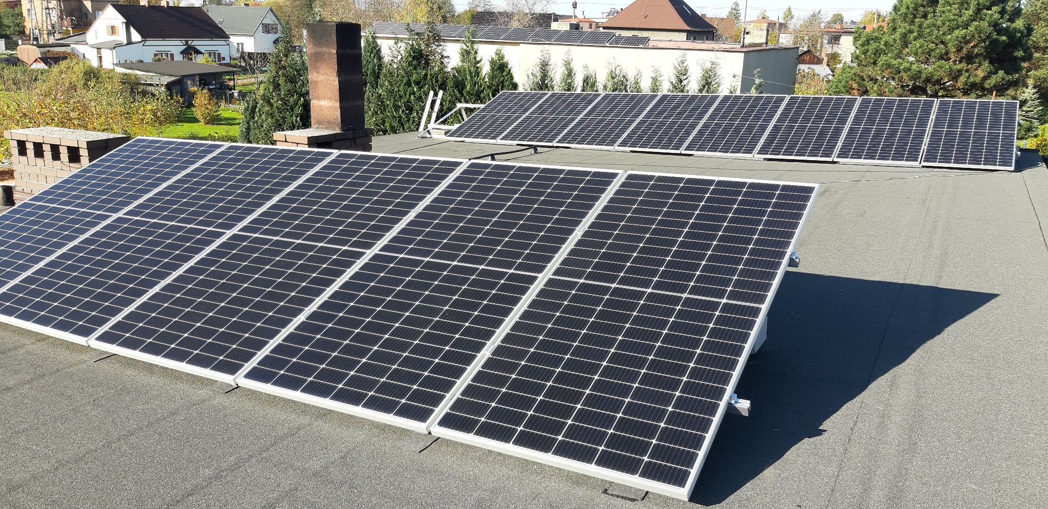 Panele słoneczne zamontowane na płaskim dachu budynku mieszkalnego, widoczne kominy i drabina oparta o dach.