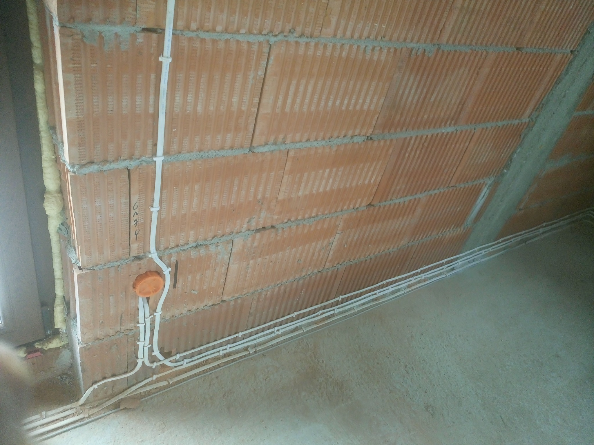 Instalacja przewodów elektrycznych w białych peszlach mocowanych do surowego muru z cegły dziurawki, widoczna pomarańczowa puszka instalacyjna i pianka montażowa wokół okna.