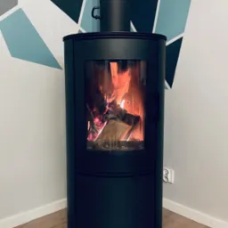 Czarny, cylindryczny kominek wolnostojący z paleniskiem, płomienie widoczne przez szybę, na podłodze zabezpieczenie z przezroczystego tworzywa, ściana z geometrycznym wzorem w tle.