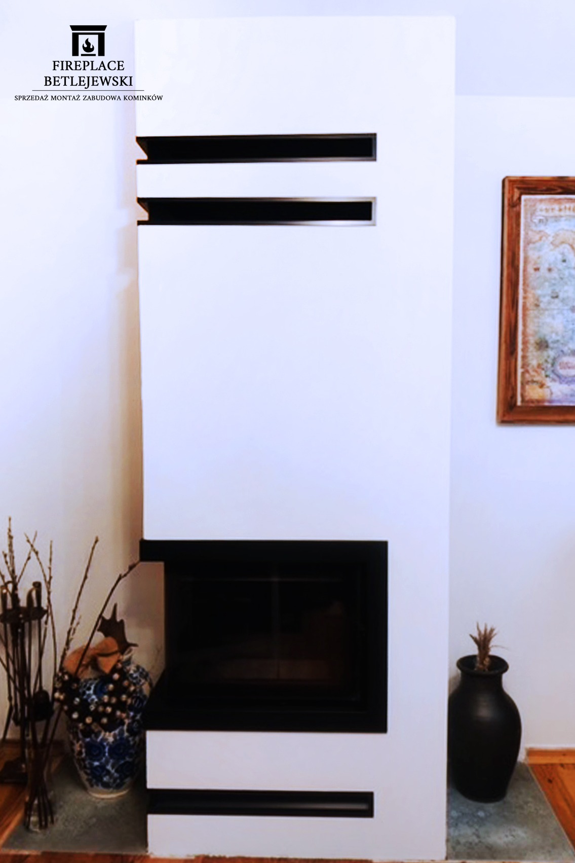 Biała zabudowa kominkowa z czarnym wkładem, dekoracyjnymi poziomymi szczelinami i wazonami po bokach, w tle rama obrazu.