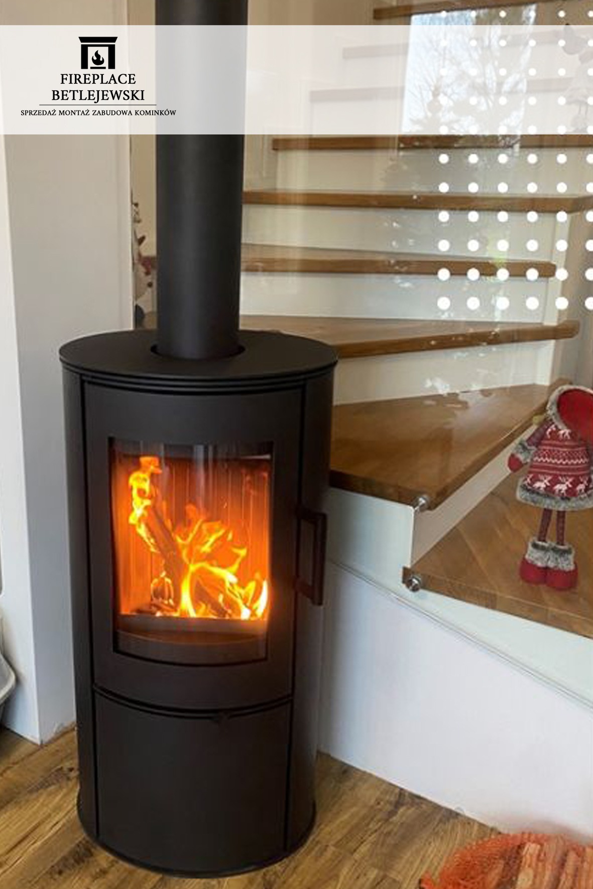 Czarny, cylindryczny piec wolnostojący z płomieniami wewnątrz paleniska, ustawiony obok drewnianych schodów z przezroczystymi panelami i dekoracyjnej figurki.