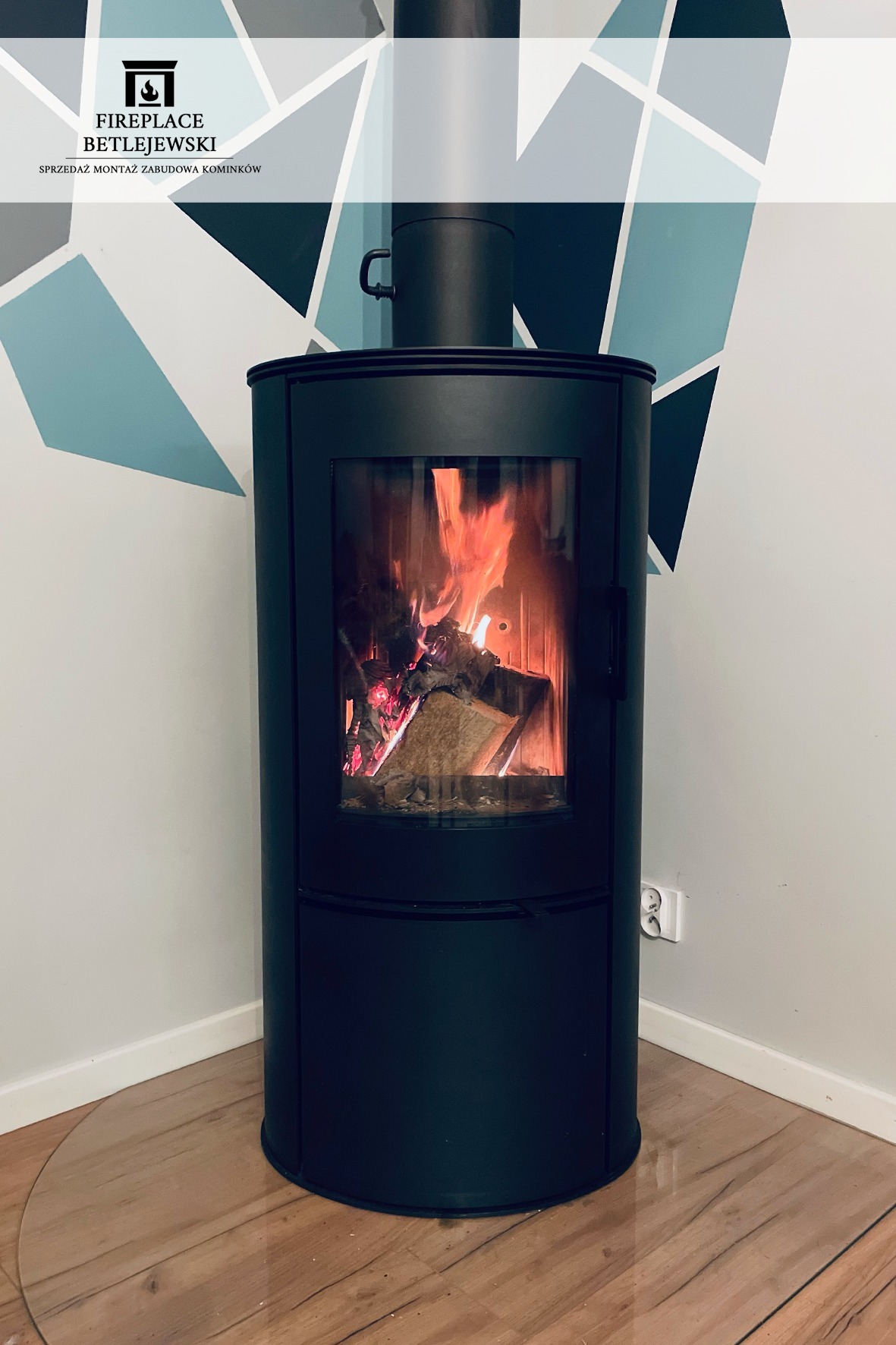 Czarny, cylindryczny kominek wolnostojący z paleniskiem, płomienie widoczne przez szybę, na podłodze zabezpieczenie z przezroczystego tworzywa, ściana z geometrycznym wzorem w tle.
