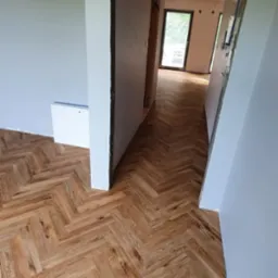 Ułożona parkietowa podłoga w jodełkę w jasnym odcieniu drewna, widok z korytarza w trakcie remontu z surowymi ościeżnicami i szarymi ścianami.