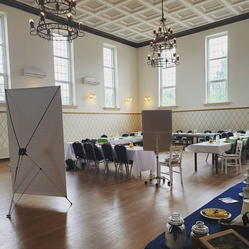 Sala konferencyjna przygotowana do wydarzenia z długimi stołami nakrytymi białymi obrusami, krzesłami, napojami oraz dwoma dużymi, pustymi tablicami reklamowymi.