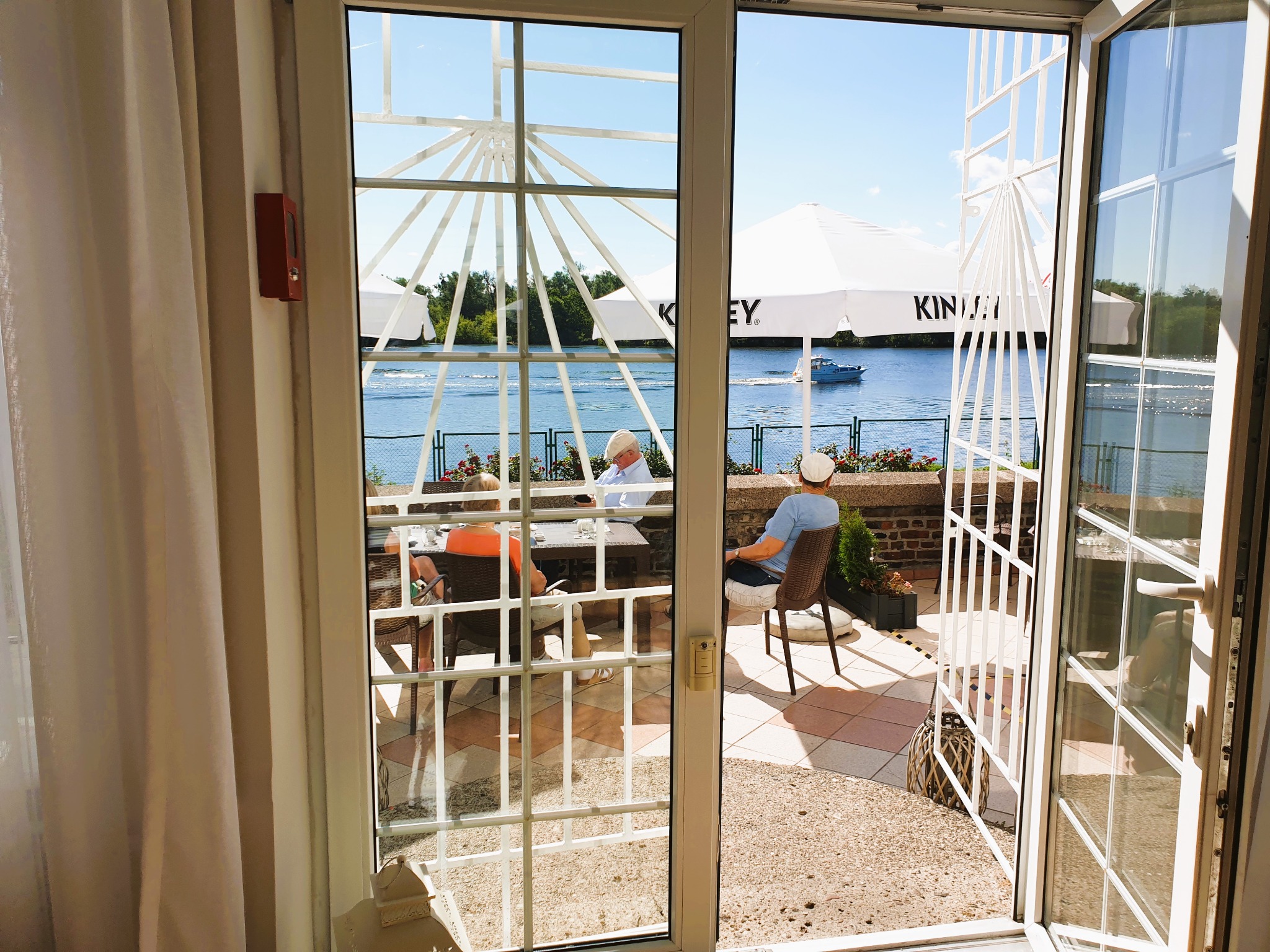 Widok przez otwarte, przeszklone drzwi na taras nad wodą z parasolami, siedzącymi osobami i przepływającą łodzią.