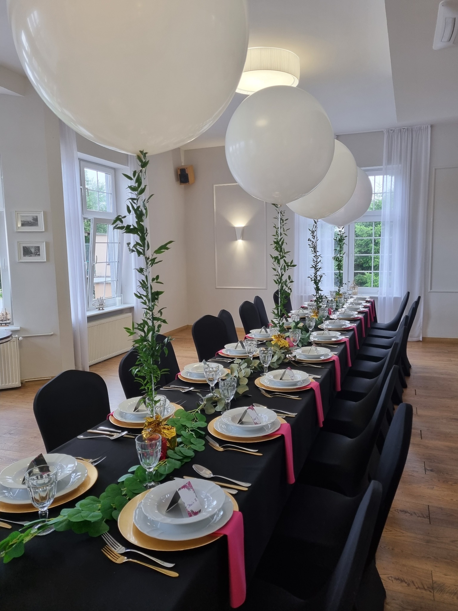 Uroczysty stół z czarnym obrusem, złotymi podkładkami, białą zastawą, dekoracjami z zielonych gałązek, różowymi akcentami i dużymi białymi balonami unoszącymi się nad stołem.