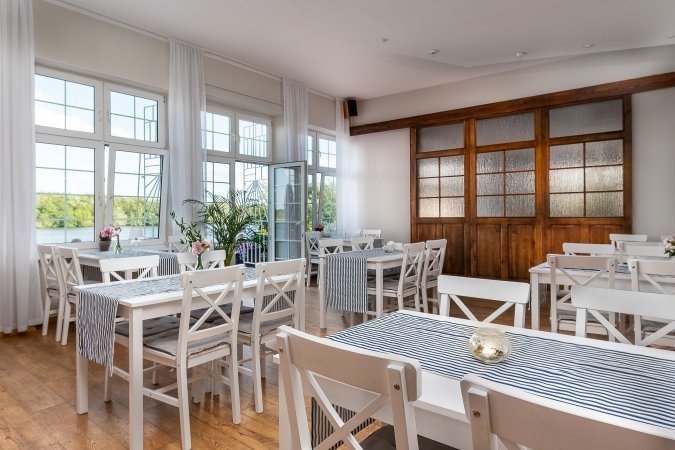 Jasne wnętrze sali restauracyjnej z białymi krzesłami i stołami nakrytymi bieżnikami w biało-niebieskie paski, widok na jezioro przez okna.