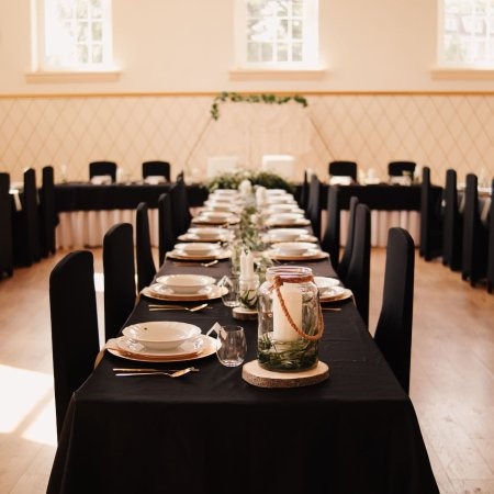 Uroczysta aranżacja stołu z czarnym obrusem, białymi naczyniami z delikatnym złotym obramowaniem, szklanymi świecznikami i dekoracją roślinną, przygotowana na przyjęcie w przestronnej sali...