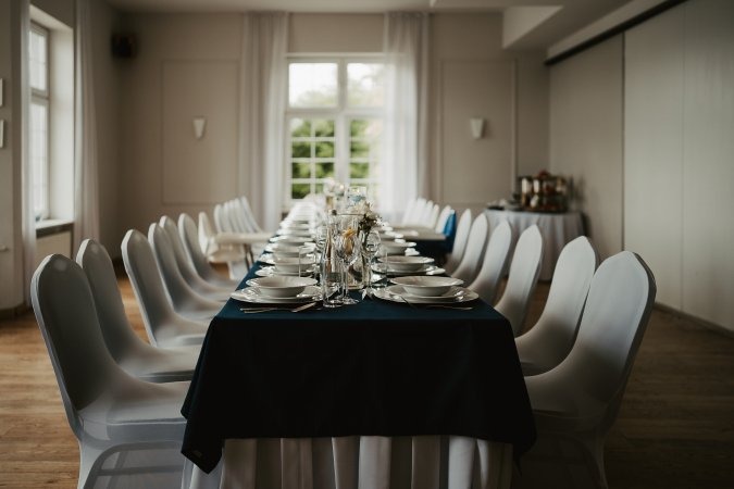 Uroczysta aranżacja stołu z ciemnoniebieskim obrusem, nakryciami i białymi pokrowcami na krzesłach w jasnym wnętrzu sali bankietowej, z widokiem na zieleń za oknem.